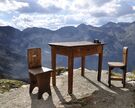 ein einfacher Holztisch und zwei Stühle in einer Berglandschaft, auf dem Tisch liegt ein Fotoapparat