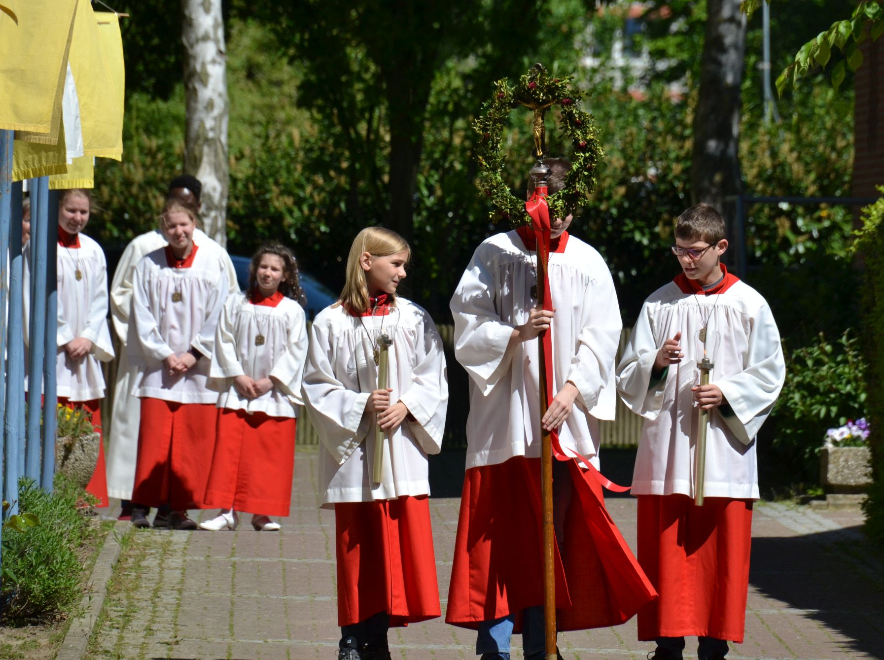Drei Messdiener mit Vortragekreuz und Flambos warten neben der Kirche auf den großen Einzug, im Hintergrund beobachten sie weitere Messdienerinnen